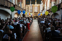 Kircheninnenraum mit gefüllten Bänken, bunter Ballonreihe entlang des Mittelgangs und einer Zeremonie auf der Bühne.
